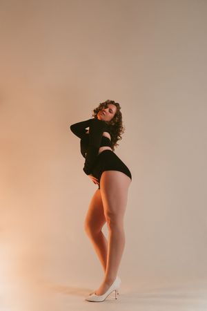 Woman in black bodysuit and white heels posing confidently against a warm beige studio background.