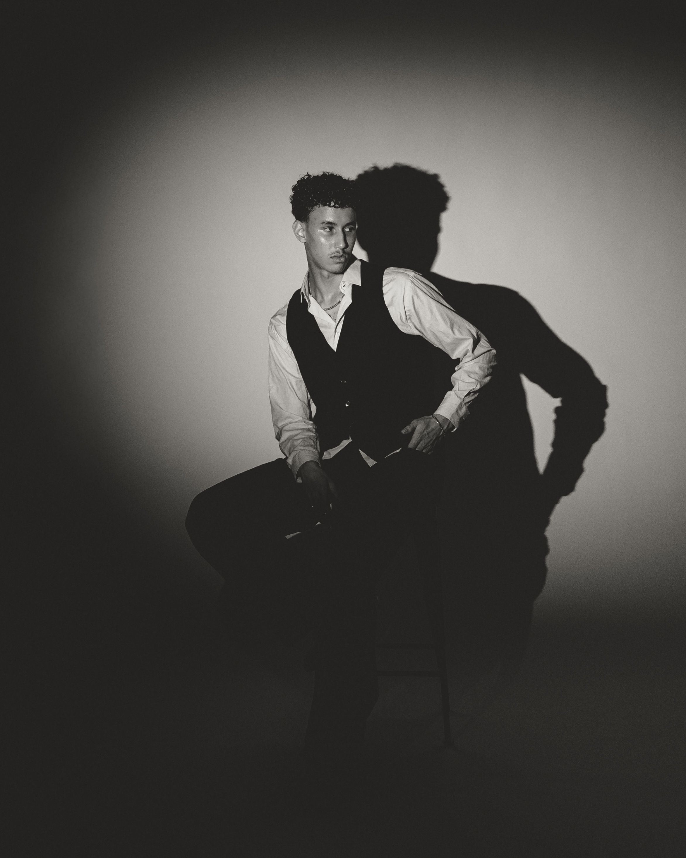 Dramatic black and white portrait with strong shadow cast against light background showing figure in vest and shirt.