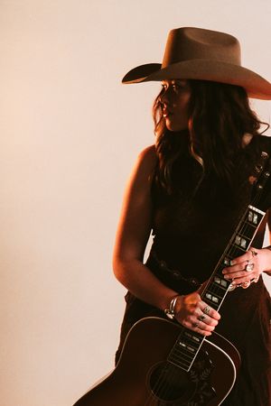 Woman in cowboy hat holding acoustic guitar, wearing black outfit, warm studio lighting, country music artist portrait.