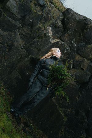 A dark-clad figure leans dramatically against a rocky cliff face covered in moss.