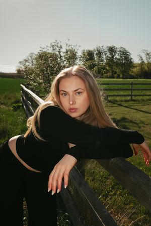 Young blonde woman in black outfit leaning on wooden fence in sunny rural field with green grass and trees.