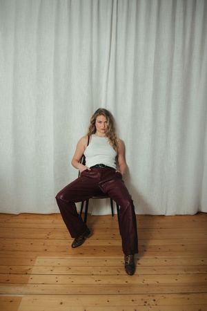 Person in white tank top and burgundy leather pants sits casually on chair against white curtain backdrop on wooden floor.