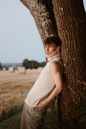 A cream knit sweater and neutral pants ensemble photographed against a large tree trunk in a field.
