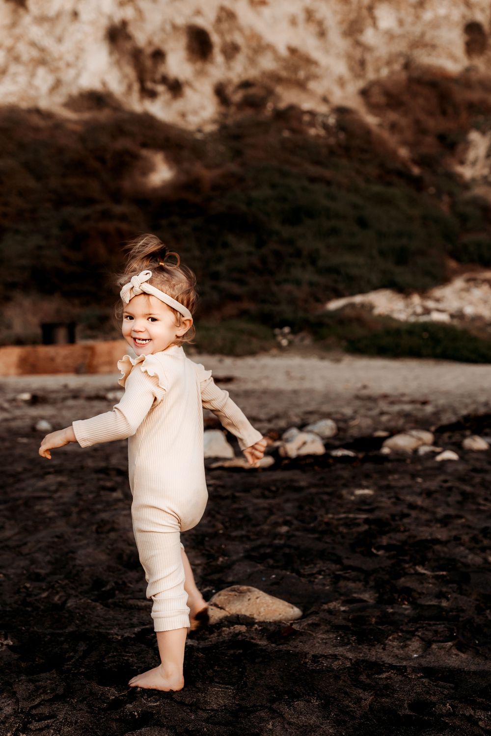 Small child in cream-colored outfit plays on dark rocky beach with mountain cliffs in background.