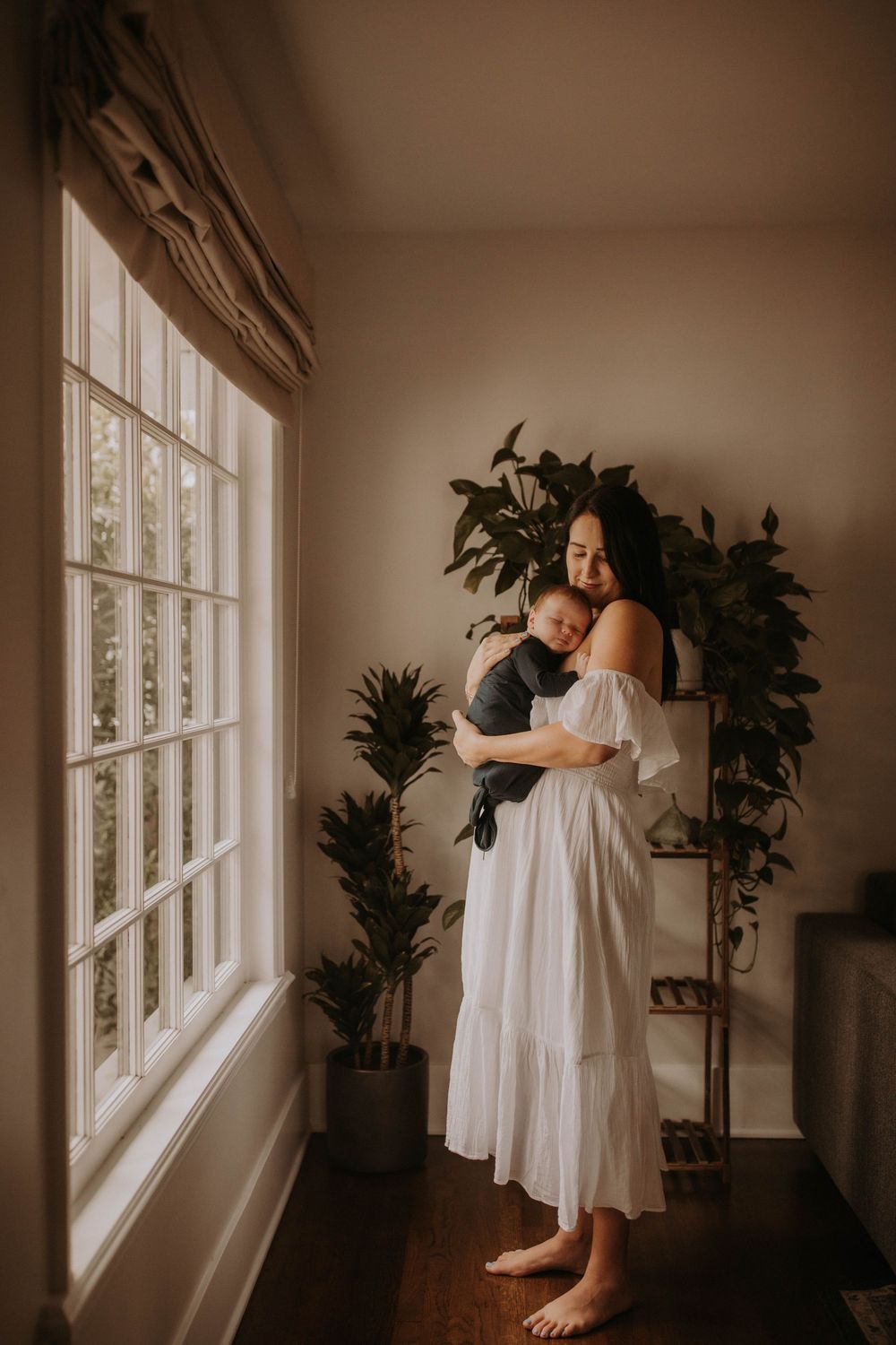 A figure in a flowing white dress dances and moves gracefully by large windows in a sunlit room with plants.