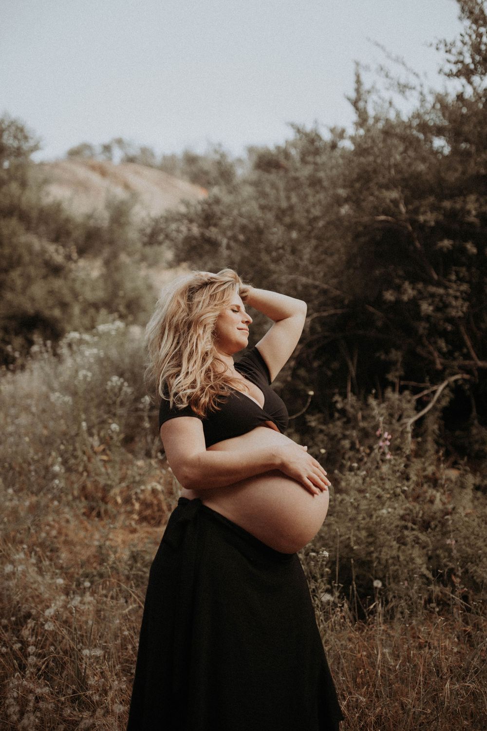 A pregnant woman in a black dress poses outdoors against a natural backdrop during golden hour.