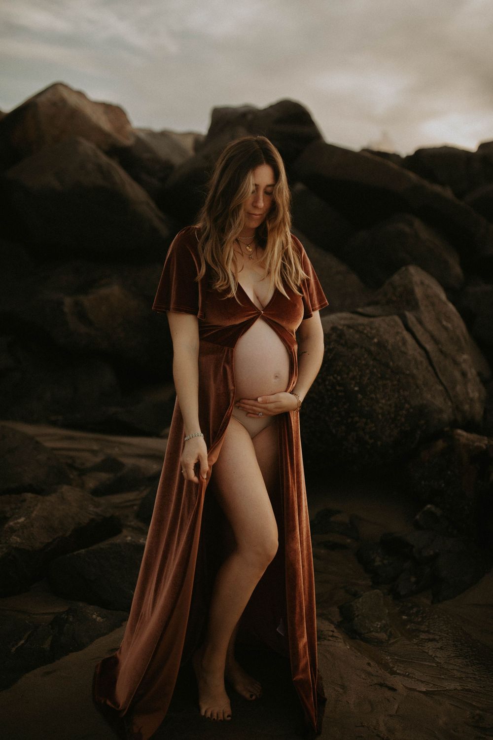 A figure in a rust-colored cardigan and beige swimsuit poses on dark rocky terrain at sunset.