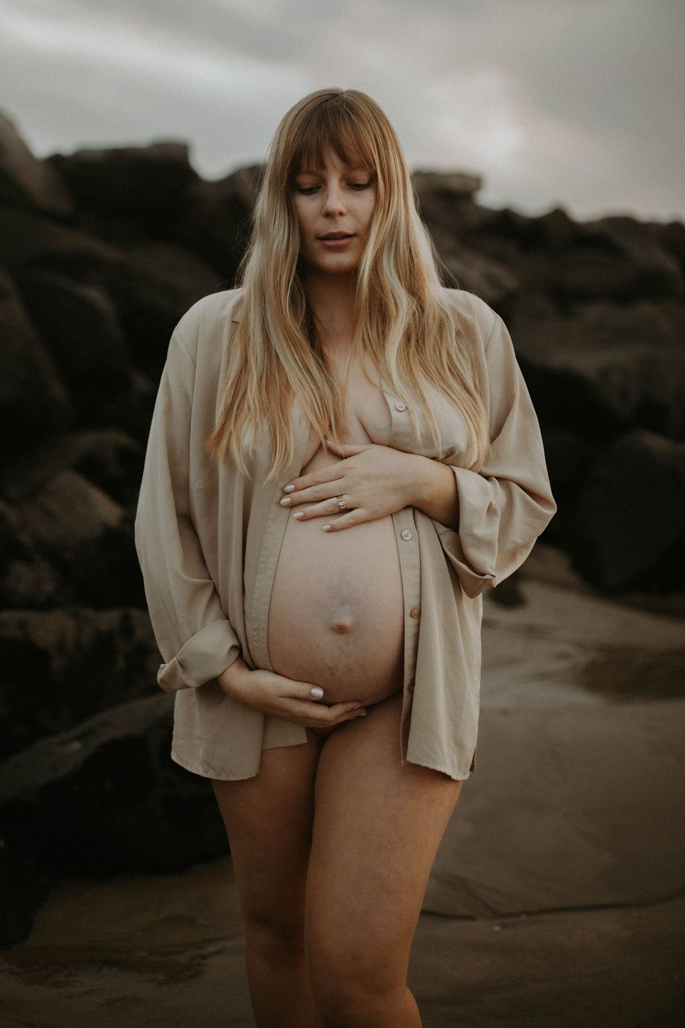 A moody maternity photoshoot features a woman in a beige shirt against a dark outdoor backdrop.