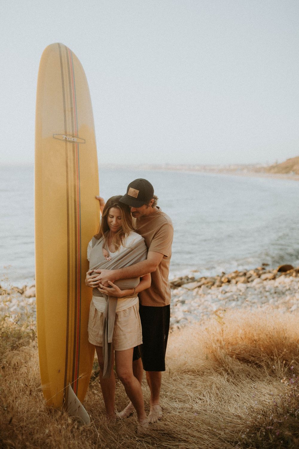 A couple embraces on a coastal hillside with a yellow surfboard leaning against dry grass at sunset.