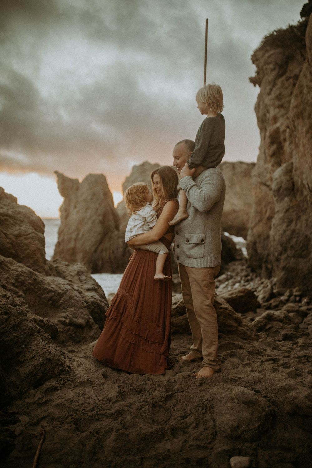 A moody coastal photoshoot at sunset shows figures standing among dramatic rocky formations by the ocean.