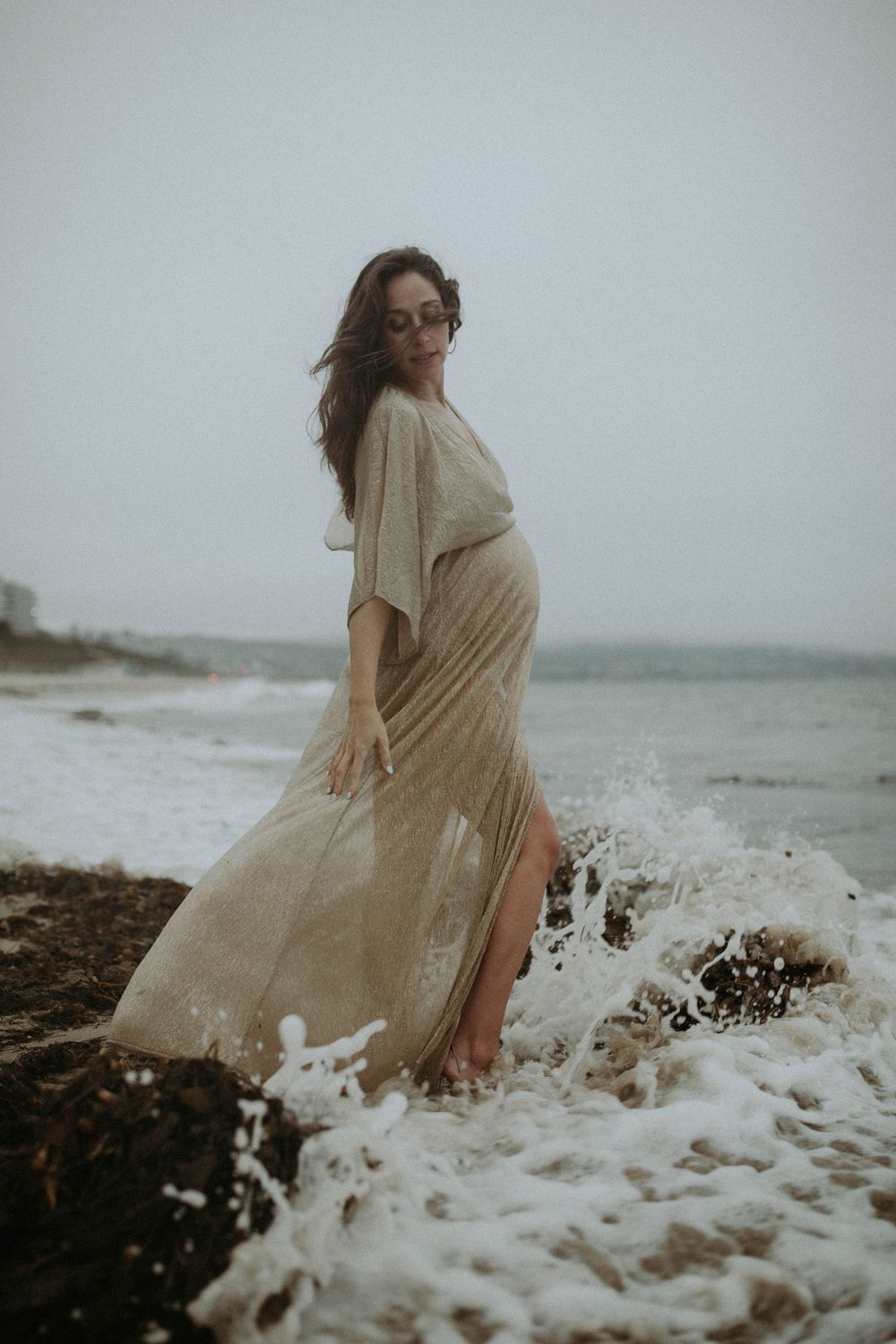 A flowing beige dress catches ocean waves splashing dramatically against rocky coastal shores on a moody overcast day.