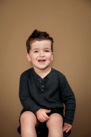 A young child in a dark sweater smiles while sitting against a brown backdrop in studio-style photographs.