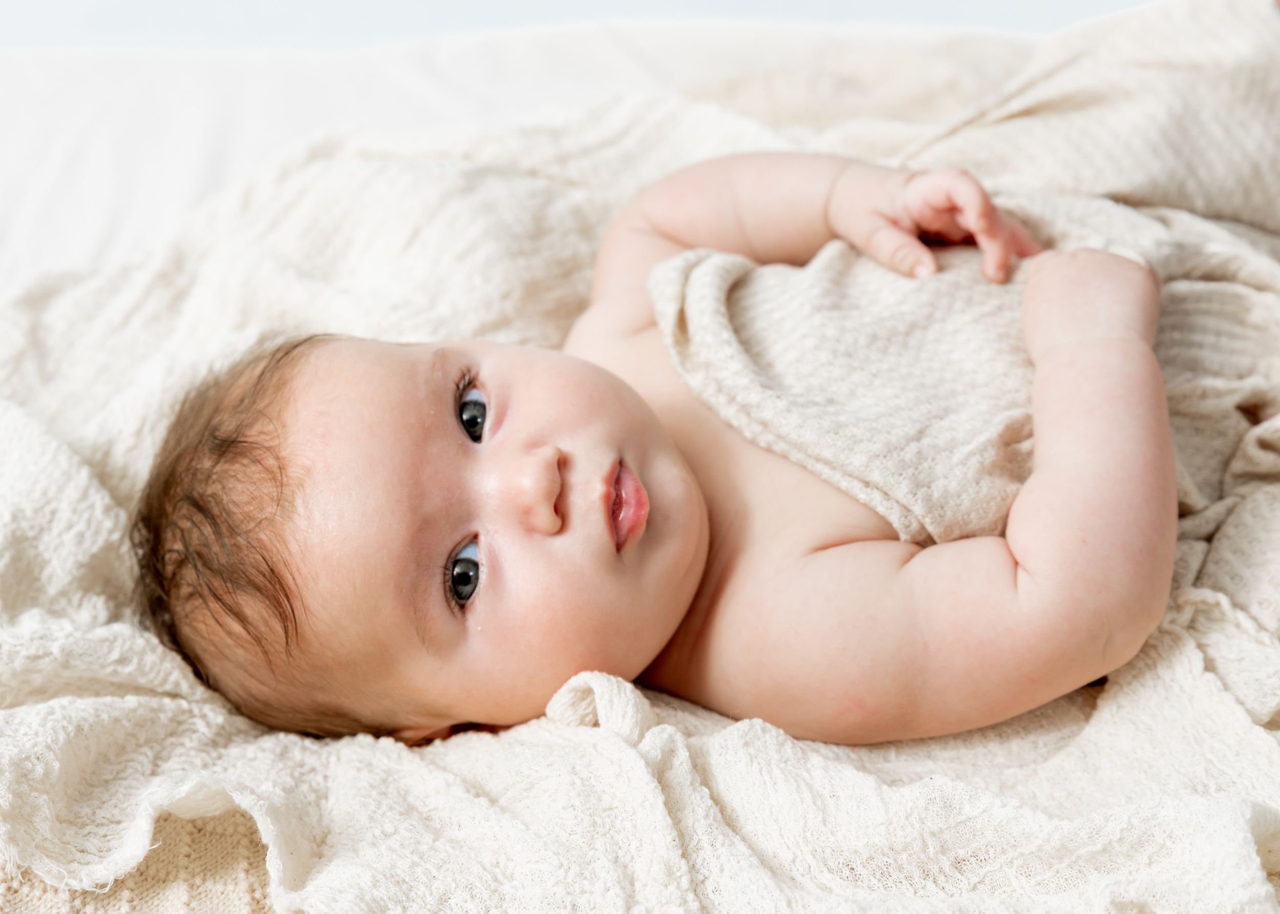 A young infant wearing white lies peacefully on a soft textured blanket, looking upwards with bright eyes.