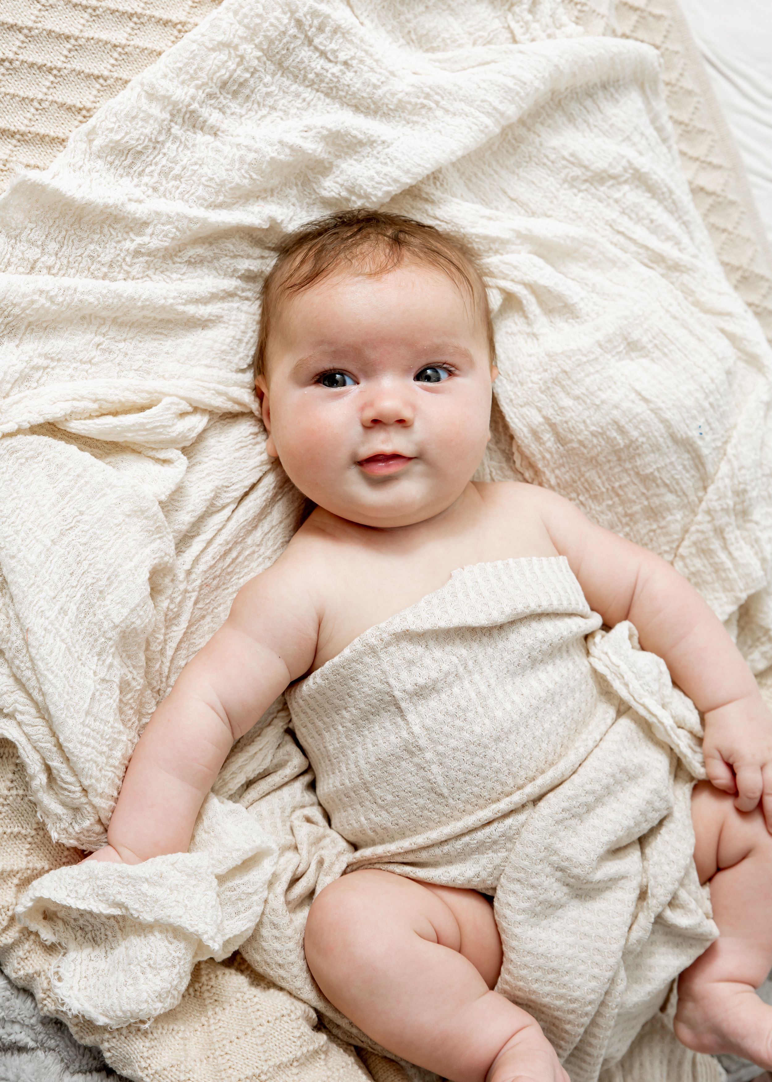 A baby wearing a white textured romper lies on white bedding while showing different poses in a sequence of photos.