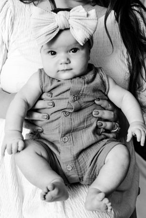 Black and white portrait of a baby wearing a romper and large white bow while being held against white fabric.