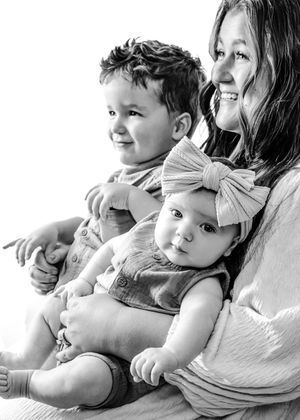 Black and white portrait of an intimate moment between siblings and mother sitting together with baby wearing large bow.