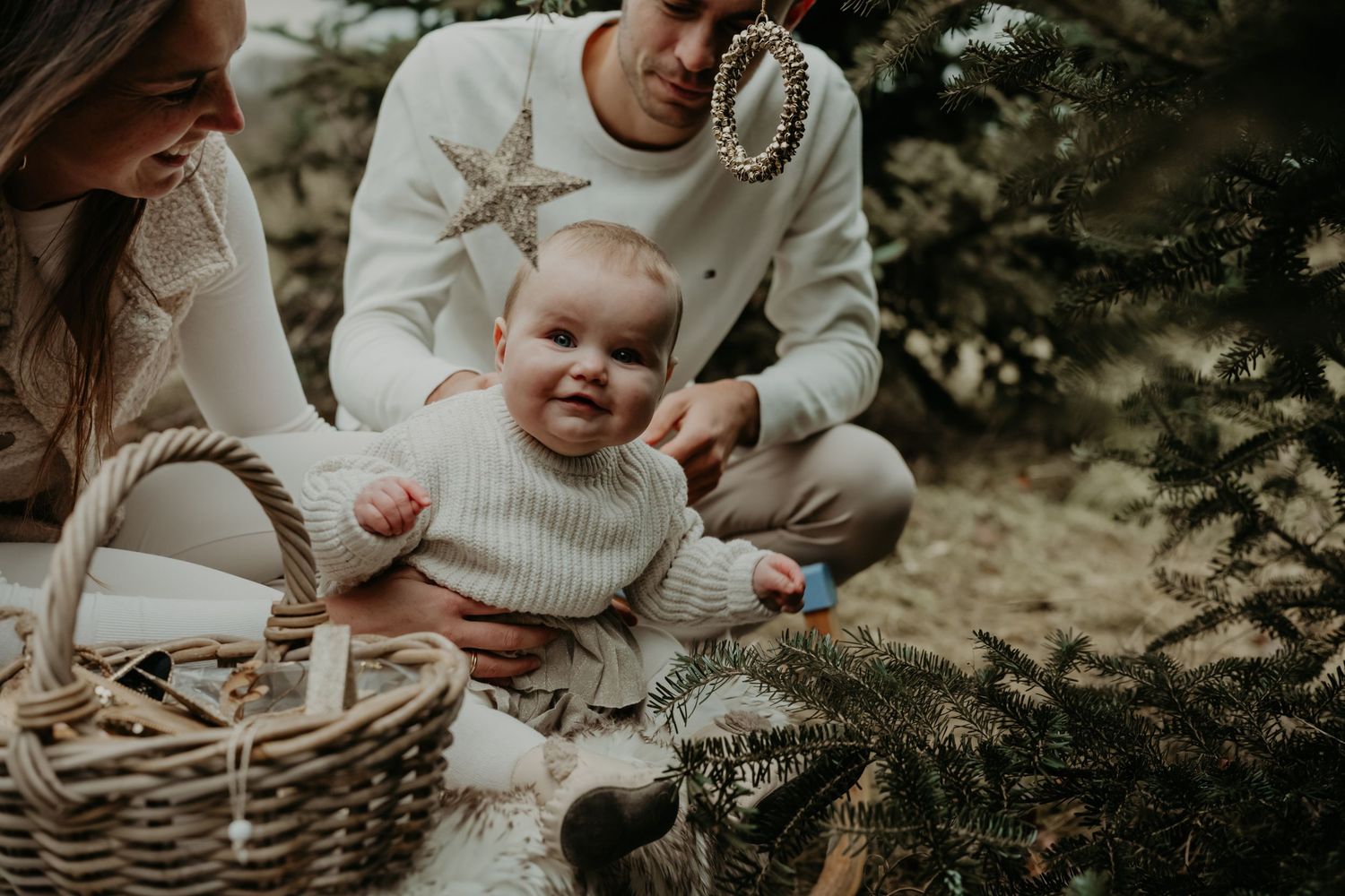 A happy baby in a white knit sweater sits outdoors with others near a woven basket during a family photo session.