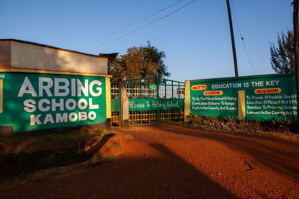 Green entrance gate and sign for Arbing School Kamobo with dirt ground and power lines overhead at sunset.
