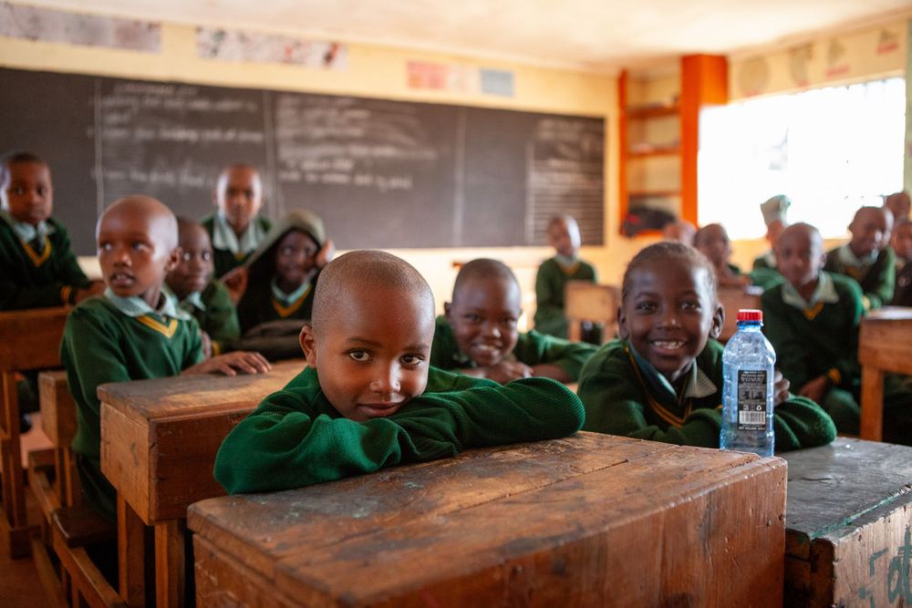 Students in green uniforms sitting attentively at wooden desks in a bright classroom setting.