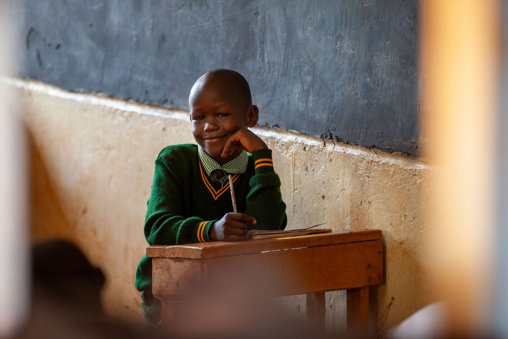 Student in green sweater sits at wooden desk in classroom with chalkboard visible in background.
