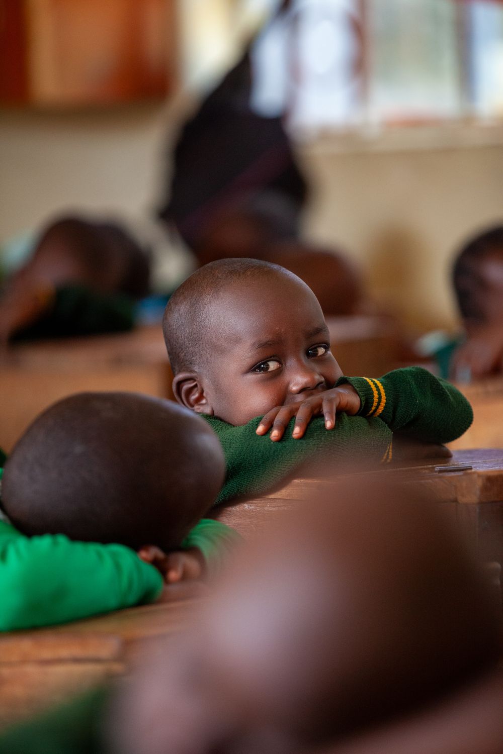 Student in green uniform rests head on desk while attending class in an African classroom.