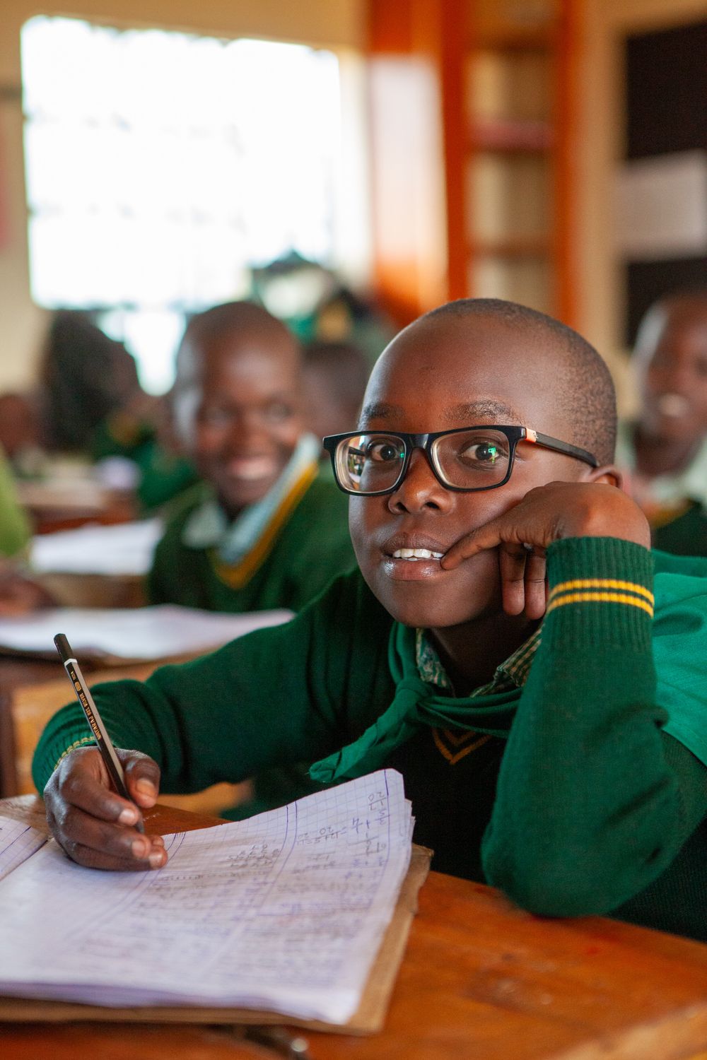 Student in green uniform and glasses sits at desk working on schoolwork while resting hand on chin.