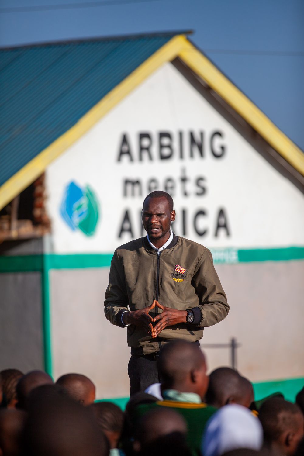 Teacher stands before students in a classroom at an African school with green buildings visible outside.