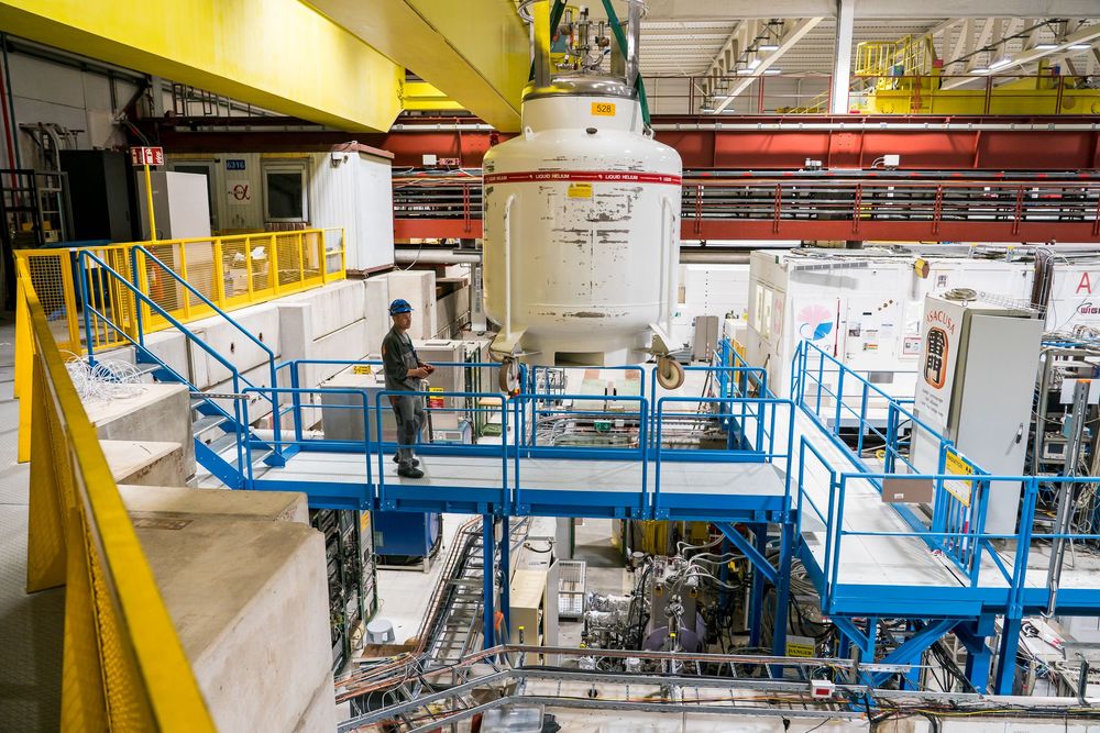 Industrial facility interior showing large scientific equipment with blue walkways and safety railings around machinery.