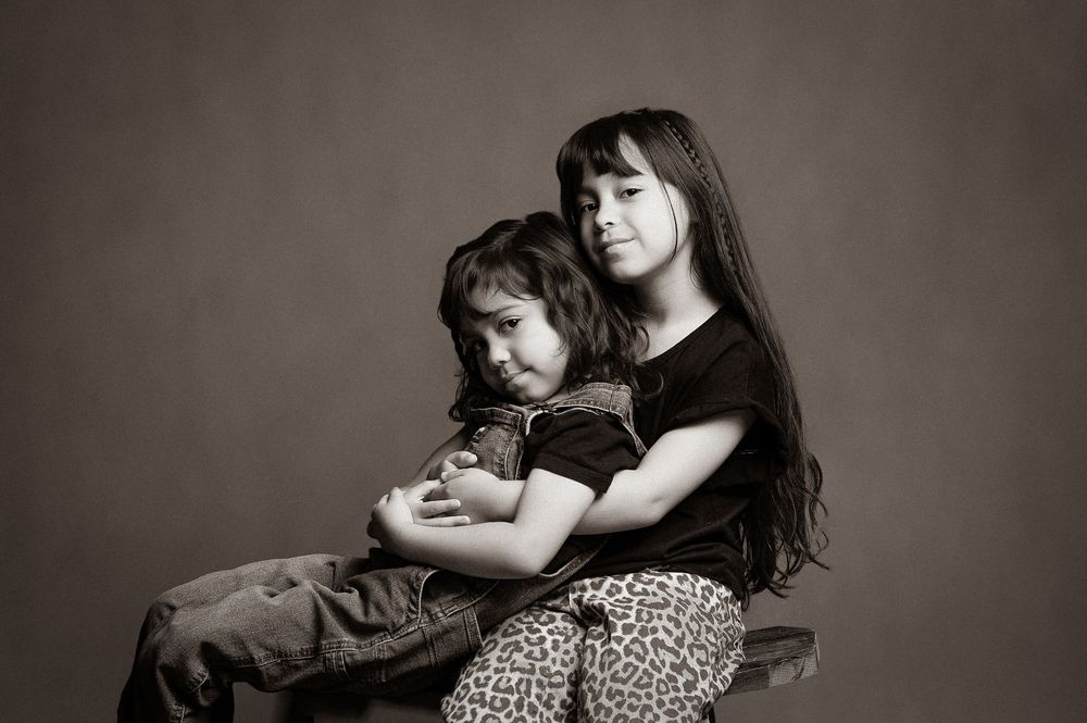 Black and white studio portrait of two siblings sitting together in casual clothing against neutral background.