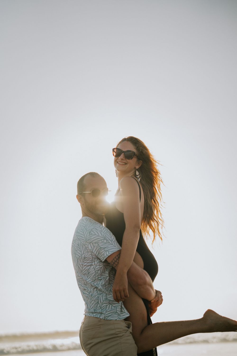 Two people share an intimate embrace on the beach with sunlight glowing behind them.