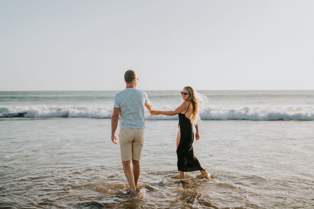 A couple walks hand in hand through gentle ocean waves at the beach.