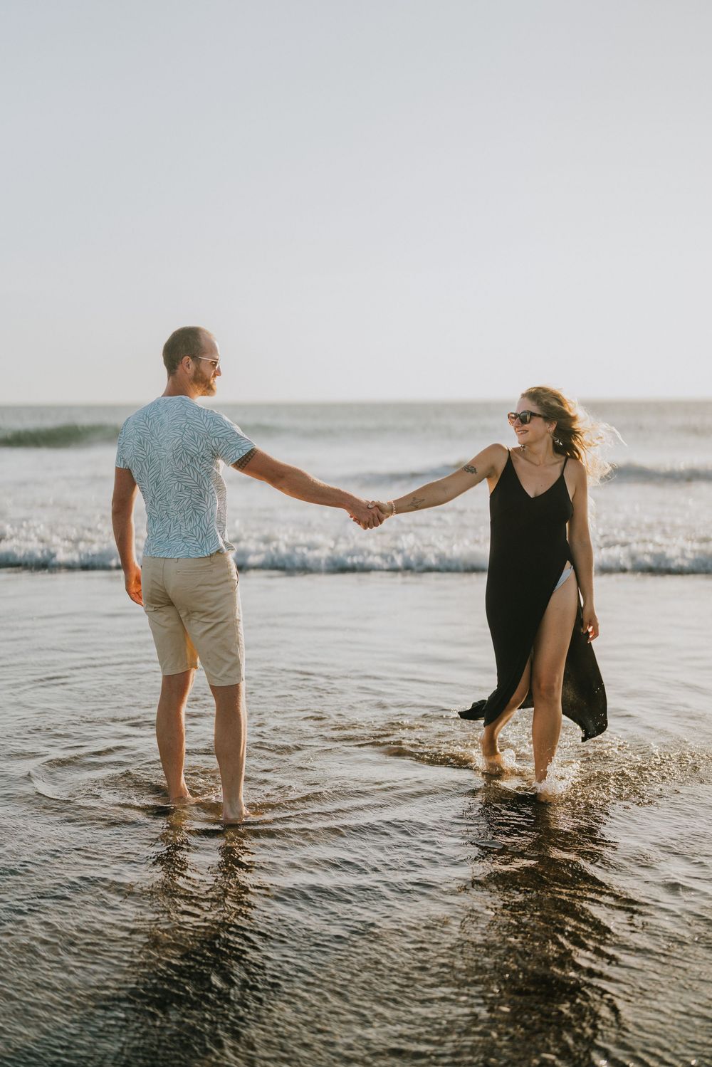 A couple plays in the shallow ocean waves at sunset creating a romantic silhouette against the water.