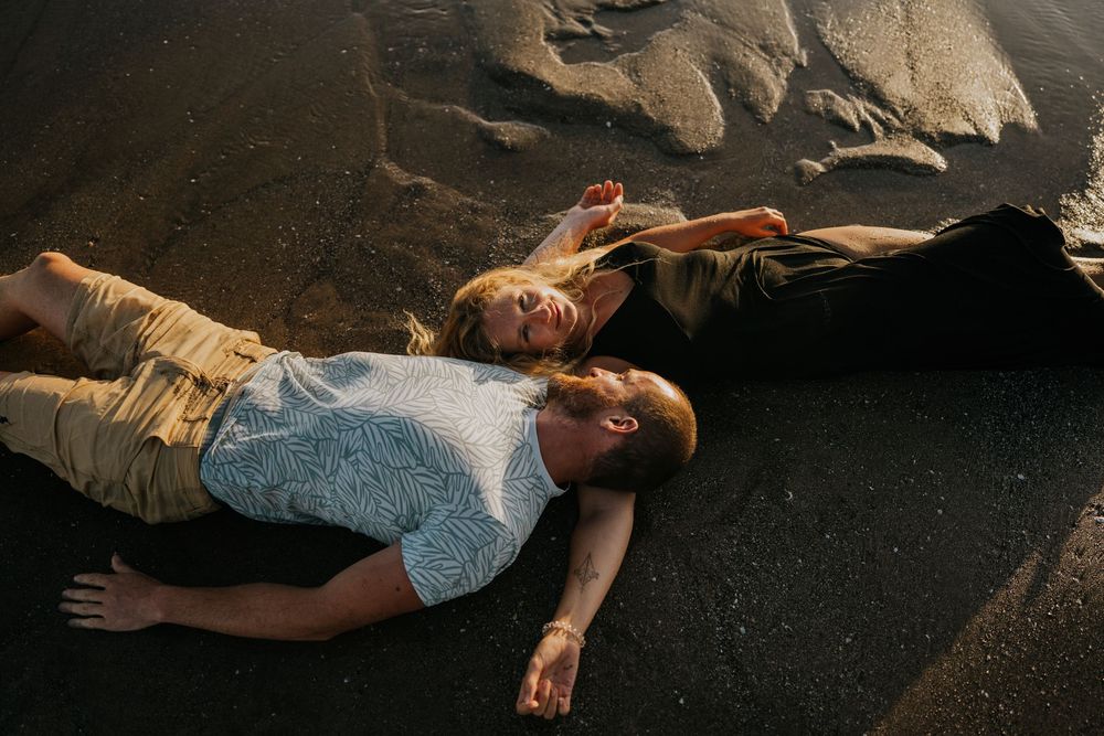 Two people lay on dark volcanic sand as ocean waves wash over them during a beach photoshoot.
