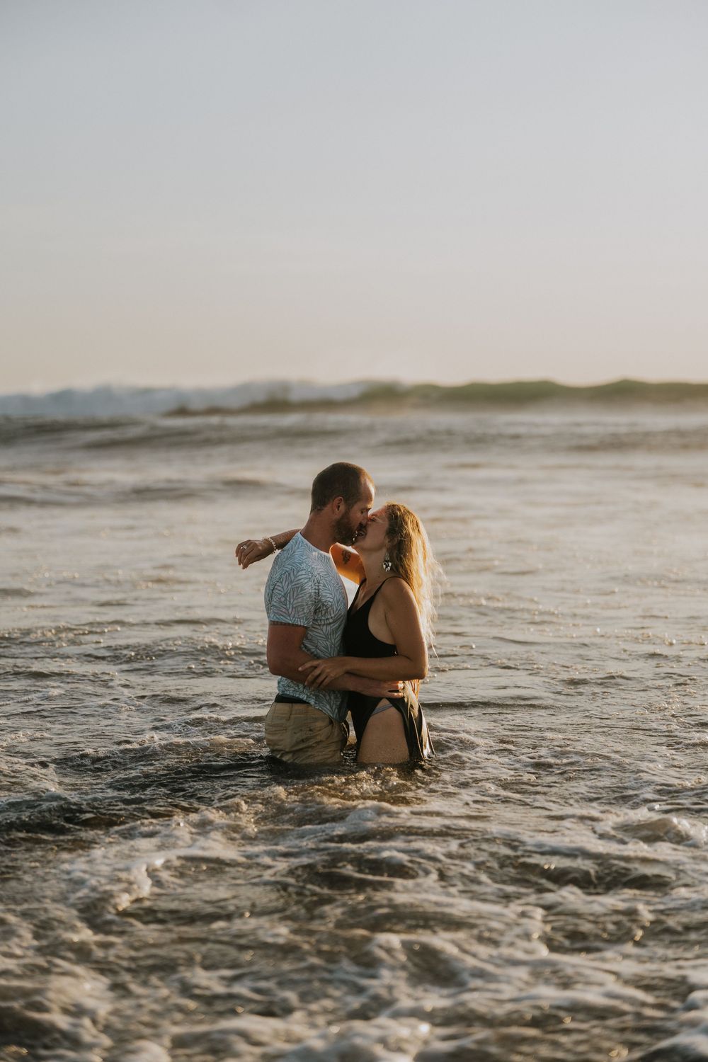 A romantic couple sits together in the ocean waves during golden hour.
