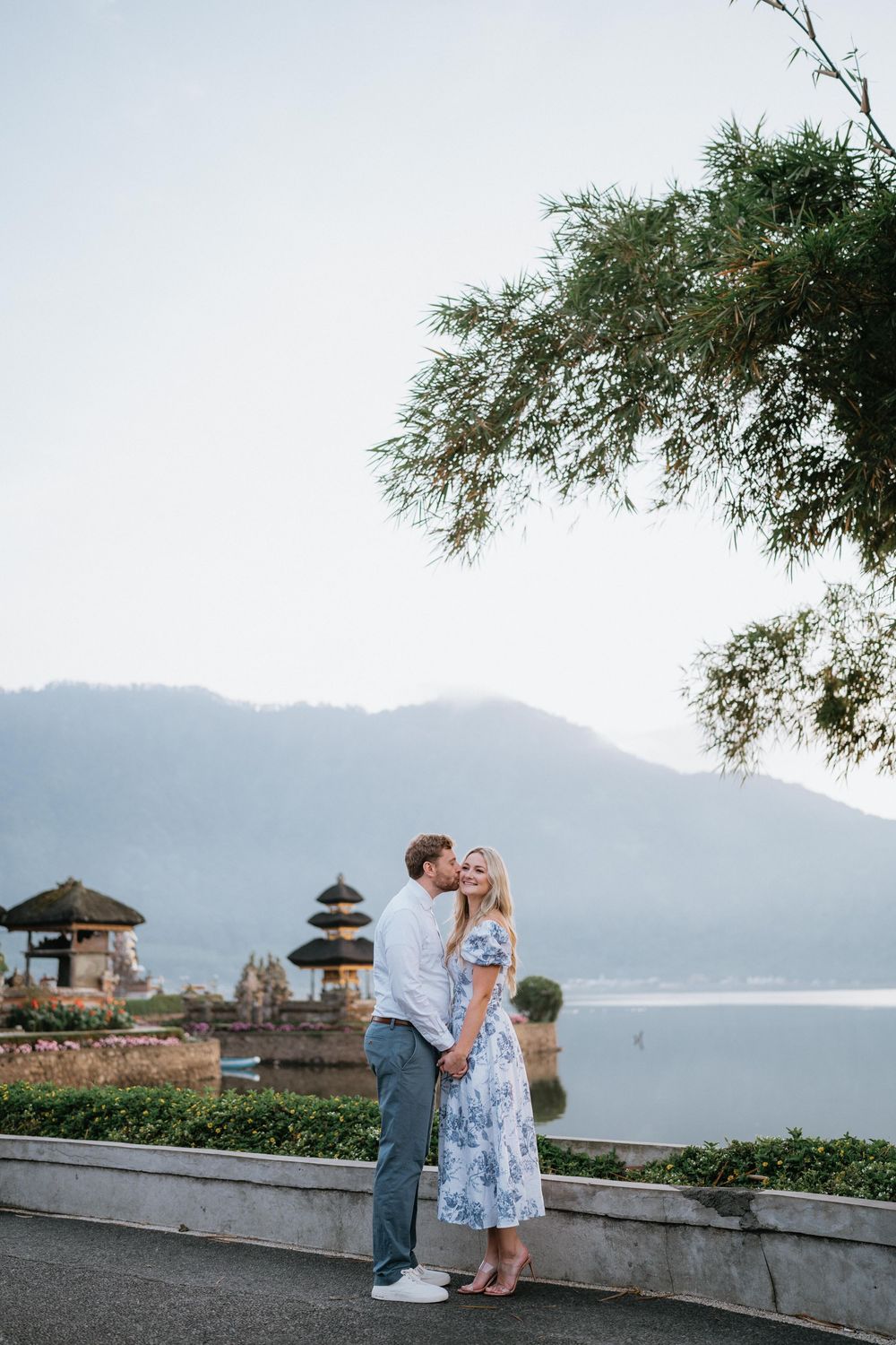 Romantic couple sharing a tender moment at a scenic temple overlooking mountains and water in Bali.