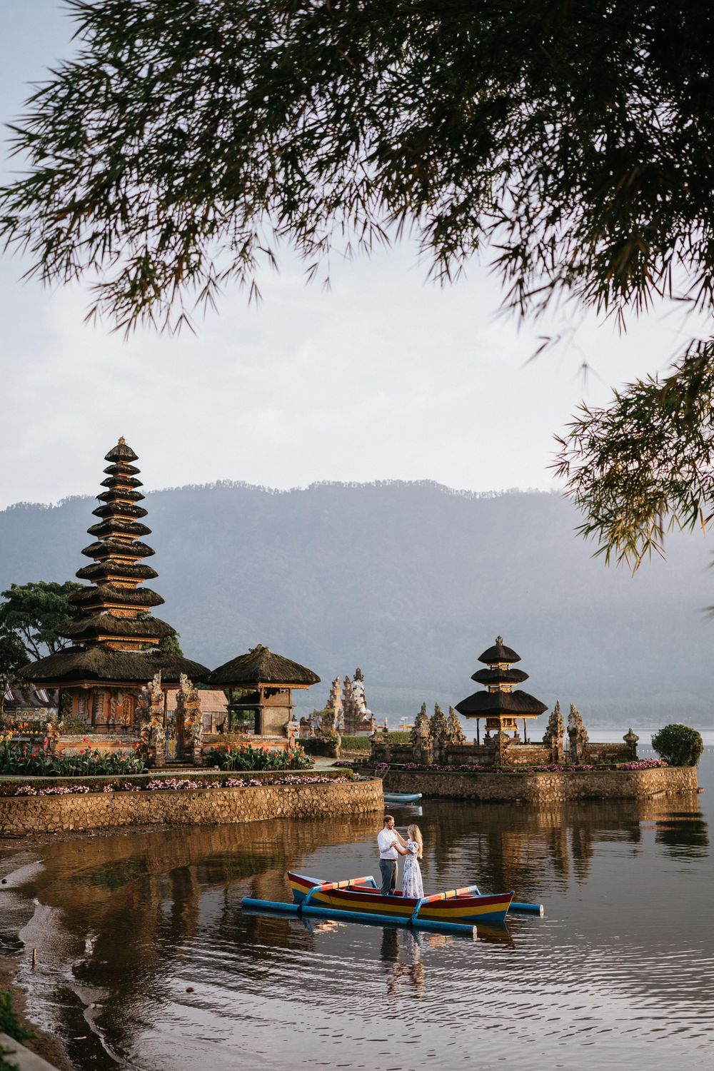 Panoramic view of Ulun Danu Beratan Temple with couple standing near traditional boat on lake.