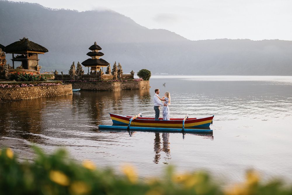 Serene lakeside view of Ulun Danu Beratan Temple with couple and boat in foreground.
