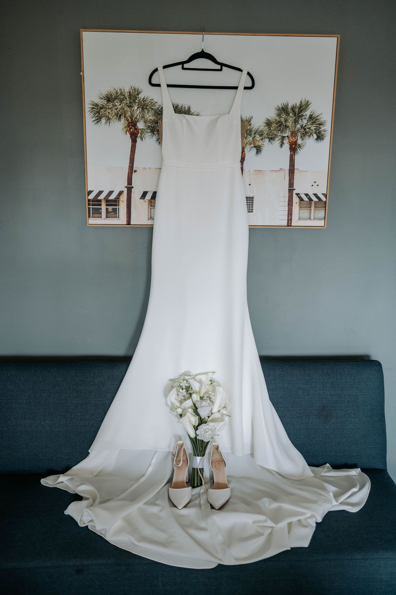 An elegant white wedding dress hanging with heels displayed underneath against a teal wall and navy couch.