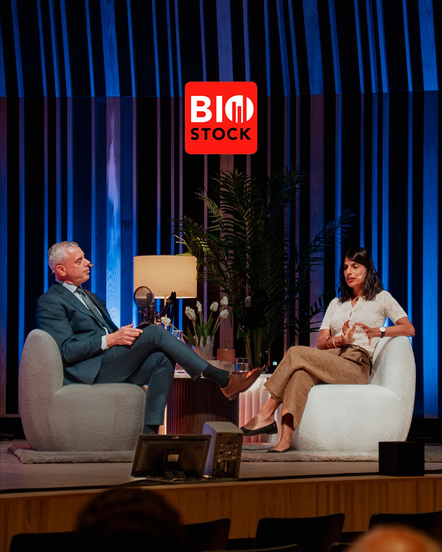 Two people engaged in conversation on stage at a Bio Stock event with blue curtain backdrop.