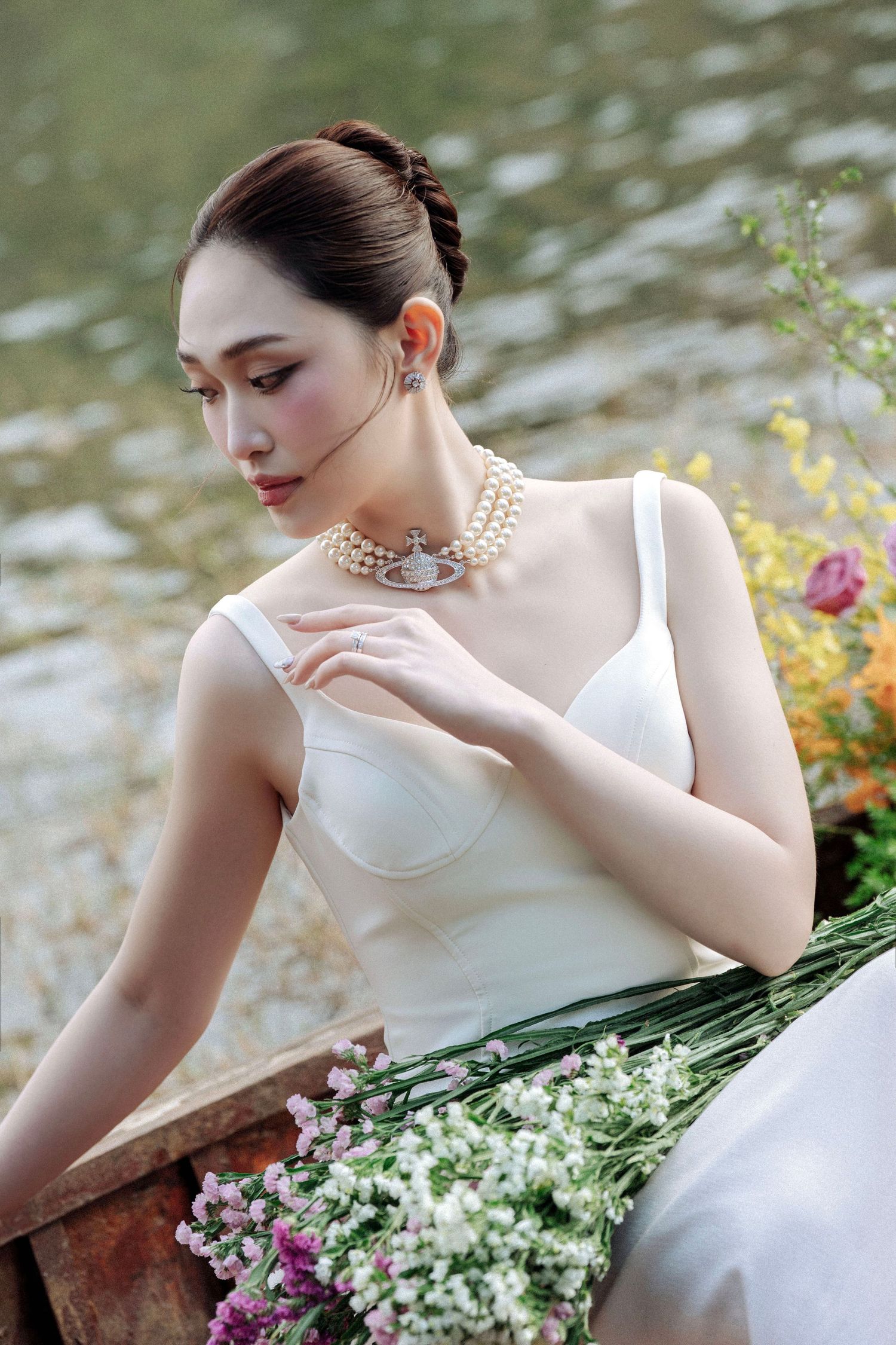 Beautiful Asian bride in white corset gown with pearl necklace holding flowers beside a lake.