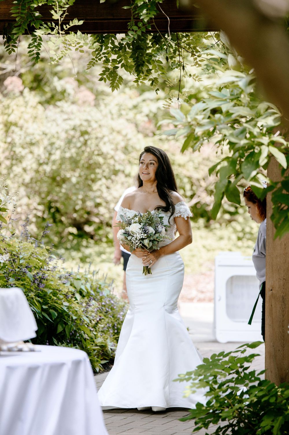 A bride in a white wedding dress stands amid lush greenery and white-draped tables at an outdoor garden venue.