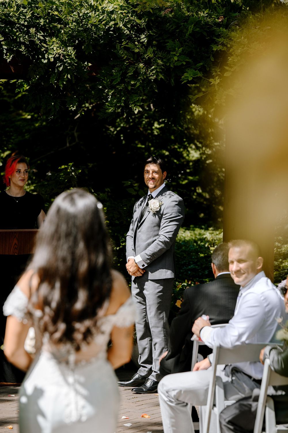 Wedding ceremony taking place outdoors with couple seated on chairs surrounded by greenery and natural light.