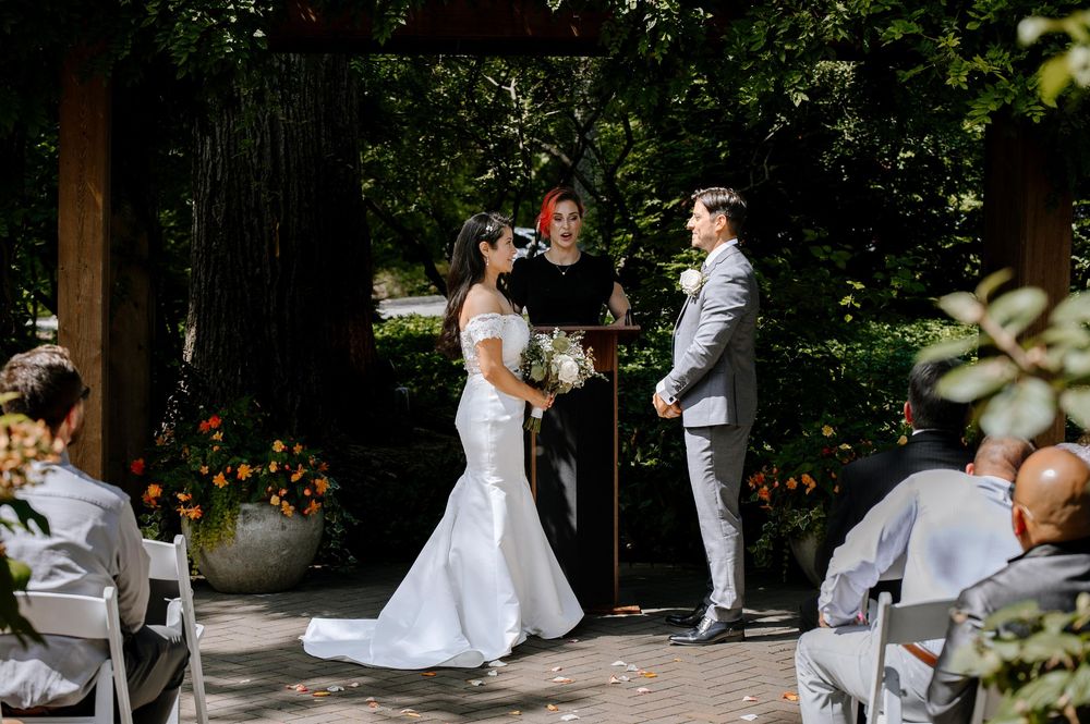 A wedding party in white attire dances together in a lush garden setting at an outdoor celebration.