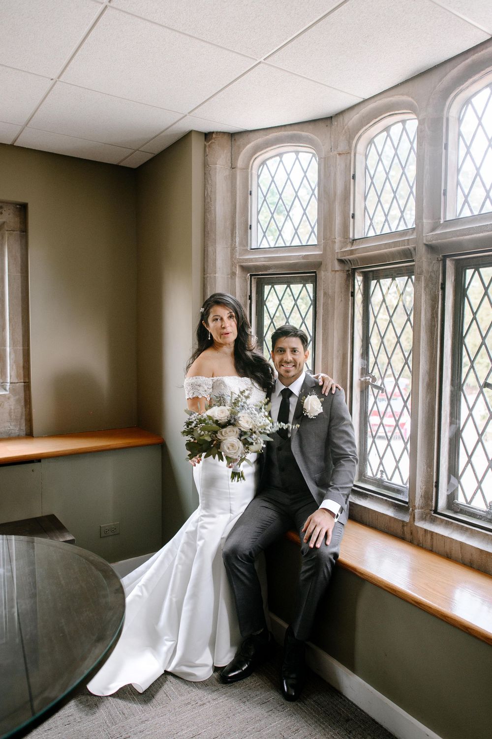 A couple in formal wedding attire pose near arched diamond-patterned windows in an elegant indoor setting.