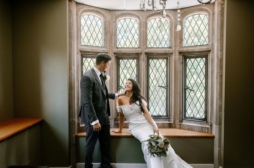 A couple shares an intimate moment together by a large diamond-paned window in a historic stone interior setting.