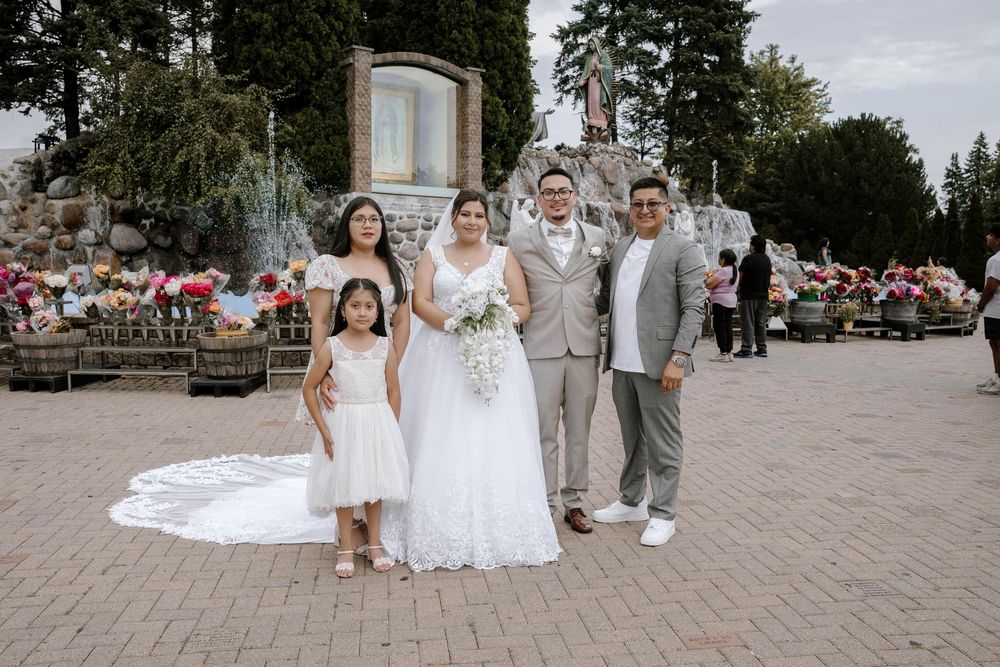 A wedding party poses outdoors on a paved area with trees and flowers in the background.