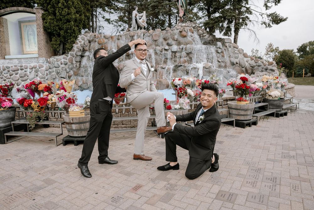 Groomsmen in formal attire pose playfully on stone patio with pink floral decorations in background.