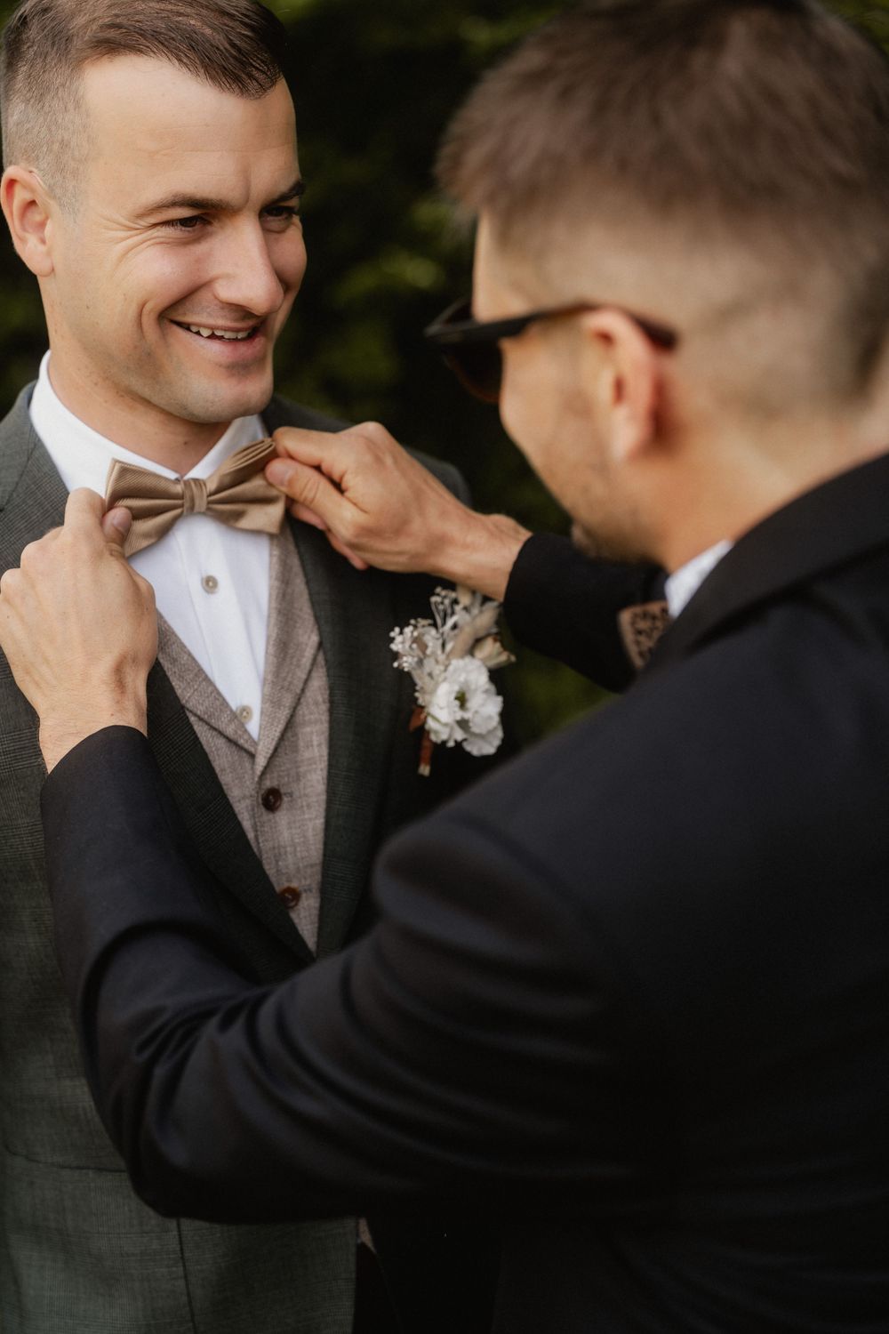 Two people in formalwear share a special wedding moment as one adjusts the other's brown bow tie.