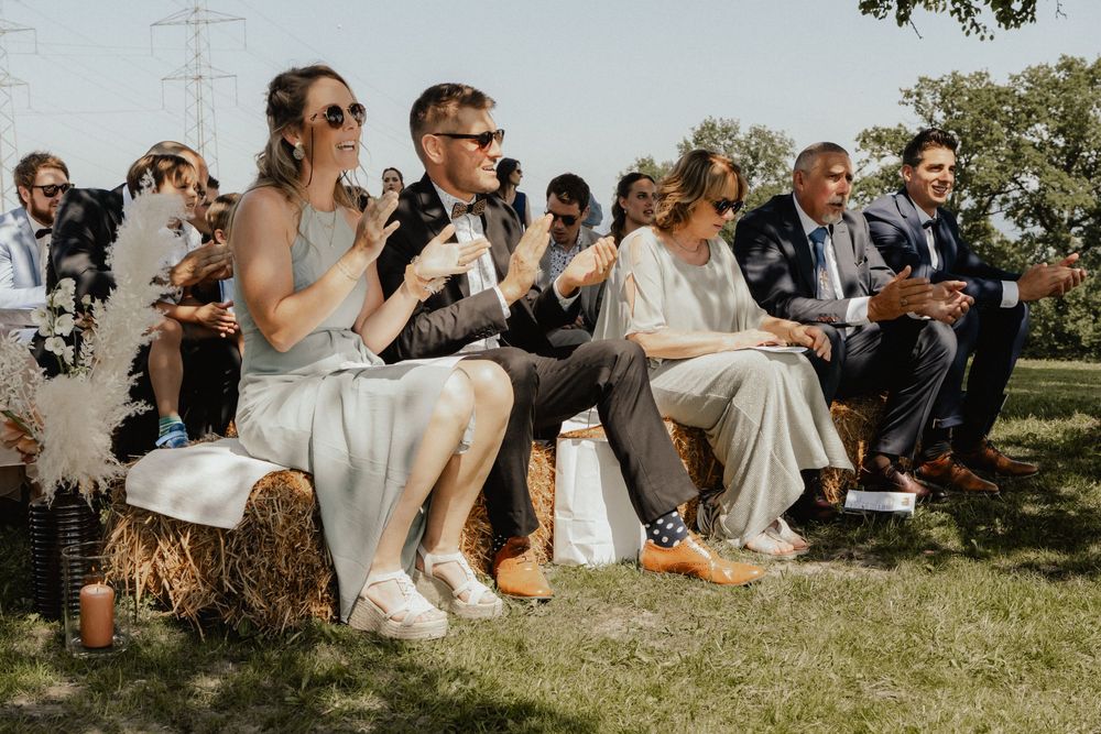 Wedding guests sit together on a grassy hillside during an outdoor ceremony in the sunshine.