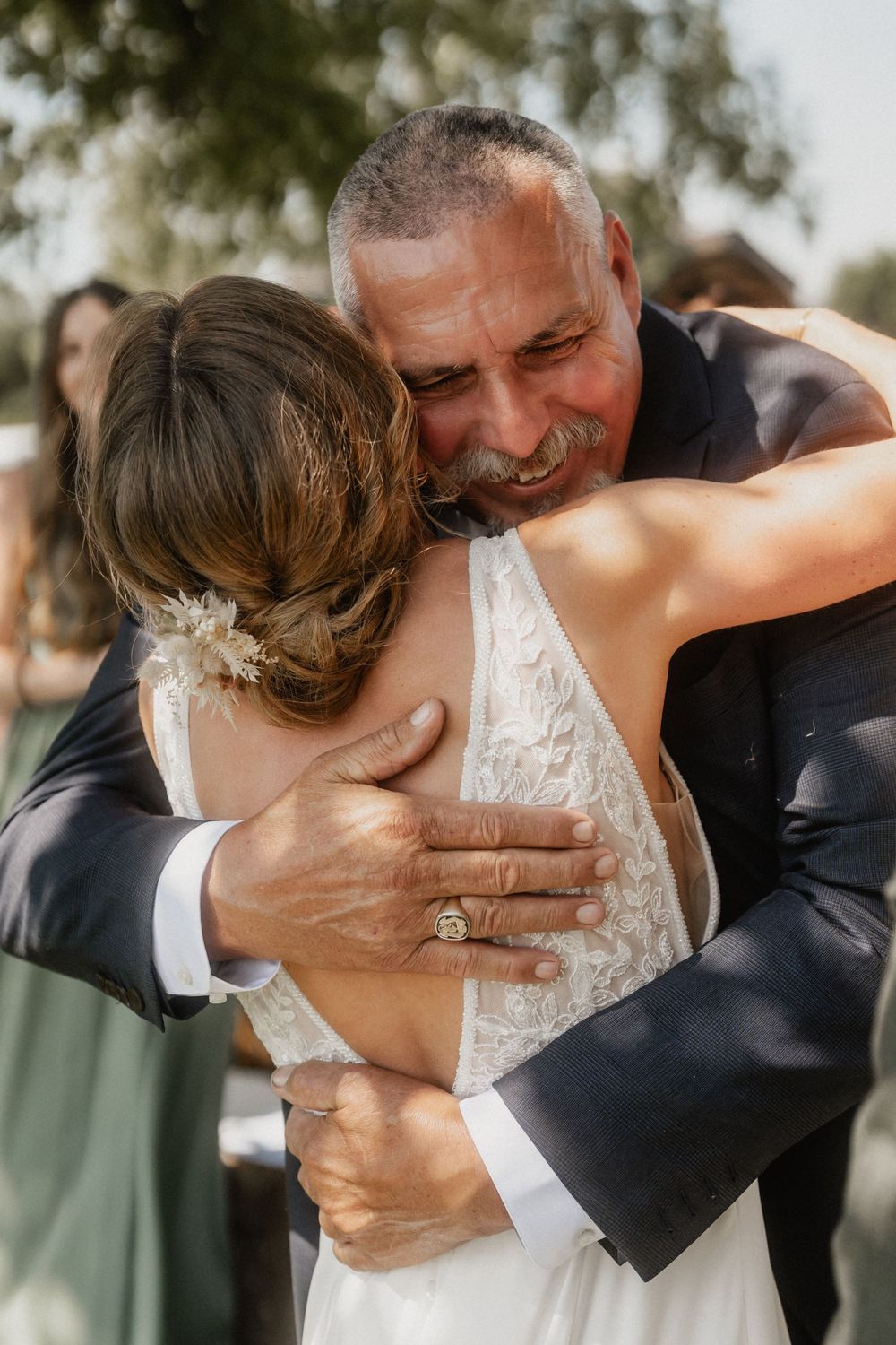 An emotional embrace between a bride in a lace dress and her loved one during an outdoor wedding celebration.